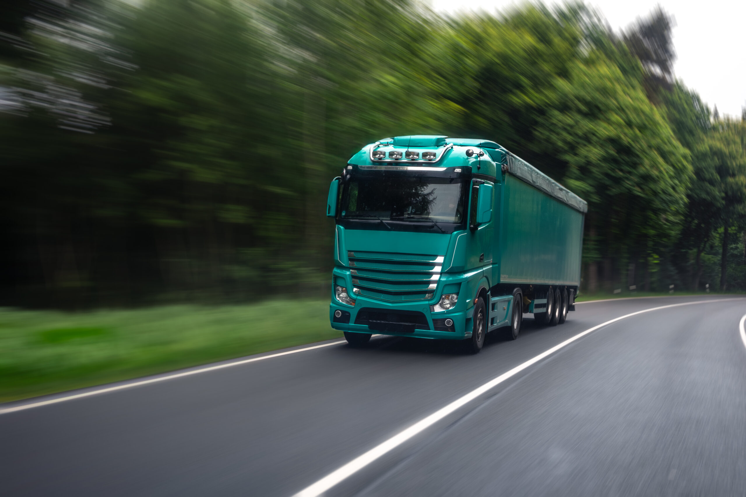 A turquoise truck drives through a forested road, illustrating the speed and power of cross-country freight delivery. The motion blur adds intensity to the logistics-themed shot.